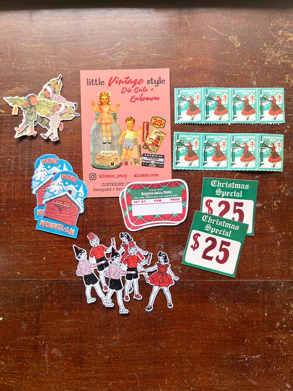 'Christmas Special' Die Cuts (18 pc) from micmoc.com at Mic Moc - A collection of vintage holiday-themed stickers and collectible ephemera arranged on a dark wooden tabletop, including a pink 'little Vintage style De-Costumizer' card, green '$25 Christmas Special' price tags, teal illustrated stamps, and colorful character stickers.